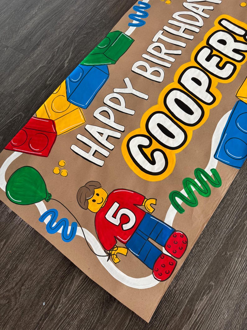 Pu&ograve; includere: Banner di compleanno dipinto a mano su carta marrone. Presenta il testo "HAPPY BIRTHAY COOPER!" in bianco con un contorno giallo. Include illustrazioni colorate di blocchi giocattolo, un palloncino e una figura con il numero 5.
