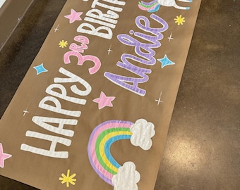 Pode incluir: Uma faixa de papel marrom com o texto "Happy 3rd Birthday Addie" em cores arco-íris. A faixa também apresenta um arco-íris, nuvens, estrelas e um unicórnio.