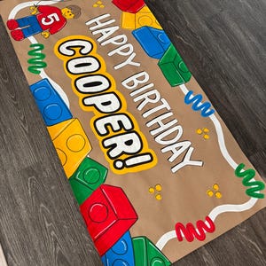 Pu&ograve; includere: Uno striscione di carta marrone con le parole "HAPPY BIRTHAY COOPER!" in lettere bianche e gialle. Lo striscione &egrave; decorato con blocchi colorati, un personaggio Lego, un palloncino verde e scarabocchi decorativi.