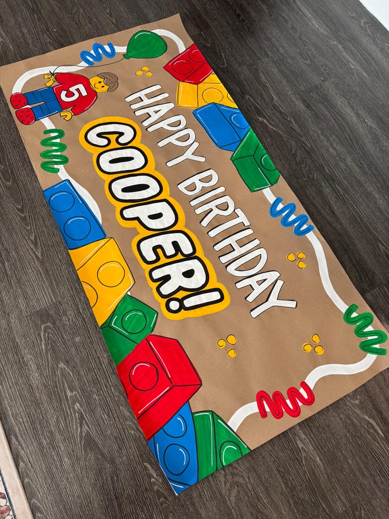 Pu&ograve; includere: Uno striscione di carta marrone con le parole "HAPPY BIRTHAY COOPER!" in lettere bianche e gialle. Lo striscione &egrave; decorato con blocchi colorati, un personaggio Lego, un palloncino verde e scarabocchi decorativi.