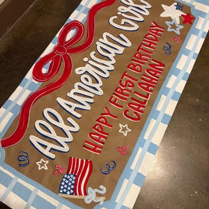 May include: A rectangular sign with the text "All American Girl" in white and blue, and "Happy First Birthday Callahan" in red. It features a red ribbon, stars, and an American flag design, all set against a brown background with a blue and white checkered border.