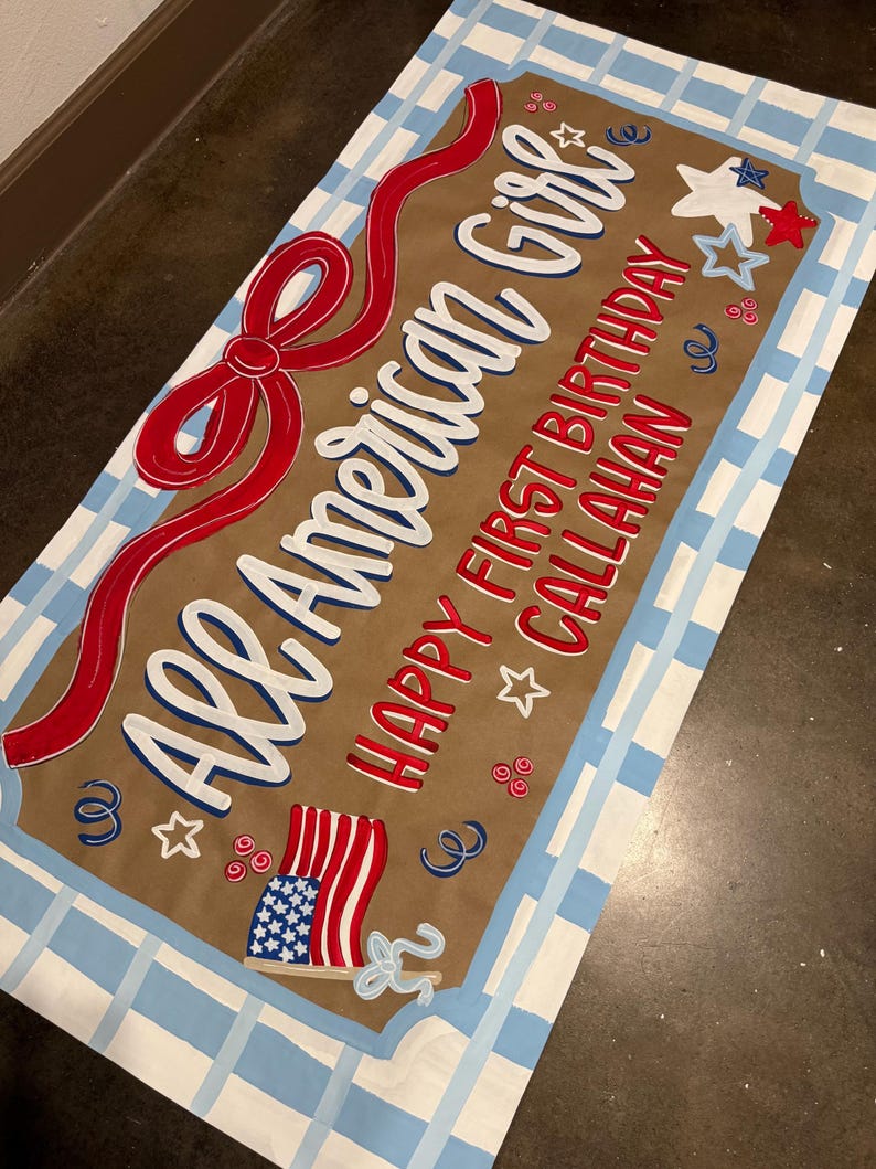 May include: A rectangular sign with the text "All American Girl" in white and blue, and "Happy First Birthday Callahan" in red. It features a red ribbon, stars, and an American flag design, all set against a brown background with a blue and white checkered border.