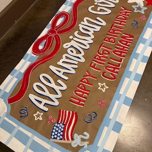 May include: A rectangular sign with the text "All American Girl" in white script, a red bow, and the words "Happy First Birthday Callahan" in red. The sign features an American flag illustration and is bordered with a blue and white plaid pattern.