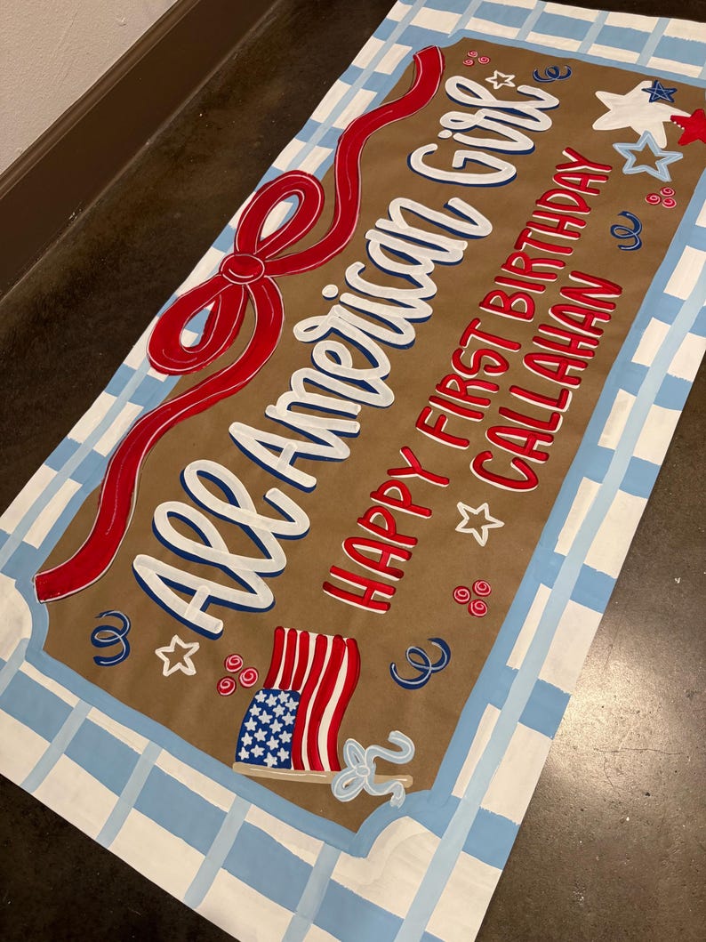 May include: A rectangular sign with the text "All American Girl" in white script, a red bow, and the words "Happy First Birthday Callahan" in red. The sign features an American flag illustration and is bordered with a blue and white plaid pattern.