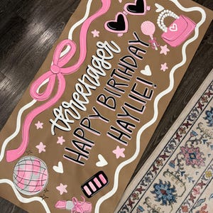 May include: A brown rectangular sign with white and pink accents. The sign says "teenager HAPPY BIRTHDAY HAYLIE!" in a handwritten font. The sign is decorated with a pink bow, heart-shaped sunglasses, a purse, stars, and other illustrations.