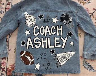 Könnte beinhalten: Hellblaue Jeansjacke mit beigem Kragen. Auf der Rückseite steht in Weiß der Schriftzug "COACH ASHLEY", begleitet von Football-Motiven wie einem Football, Helm und Sternen. Die Jacke hat lange Ärmel.