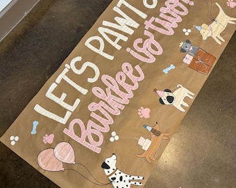 Puede incluir: Una pancarta de papel marrón con el texto "Let's Pawty! Barklee is two!" en letras blancas y rosas. La pancarta está decorada con huellas de patas, huesos, globos e ilustraciones de perros.