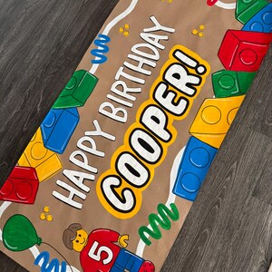 Pu&ograve; includere: Uno striscione di carta marrone con le parole "HAPPY BIRTHDAY COOPER!" in lettere bianche e gialle. Lo striscione presenta illustrazioni di blocchi colorati, una figura Lego e un palloncino verde. La figura Lego ha una camicia rossa con il numero 5.