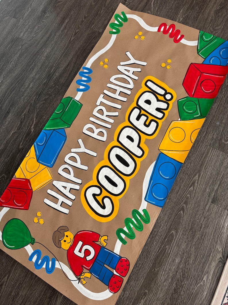 Pu&ograve; includere: Uno striscione di carta marrone con le parole "HAPPY BIRTHDAY COOPER!" in lettere bianche e gialle. Lo striscione presenta illustrazioni di blocchi colorati, una figura Lego e un palloncino verde. La figura Lego ha una camicia rossa con il numero 5.