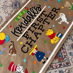 May include: A brown paper banner with the text "Kendall's FIRST CHAPTER" and colorful illustrations of storybook characters. The banner features a white border and colorful polka dots.