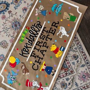 May include: A brown paper banner with the text "Kendall's FIRST CHAPTER" in black and white. The banner features colorful illustrations of storybook characters, including a mouse, a caterpillar, and Winnie the Pooh. The banner is trimmed with a white border.