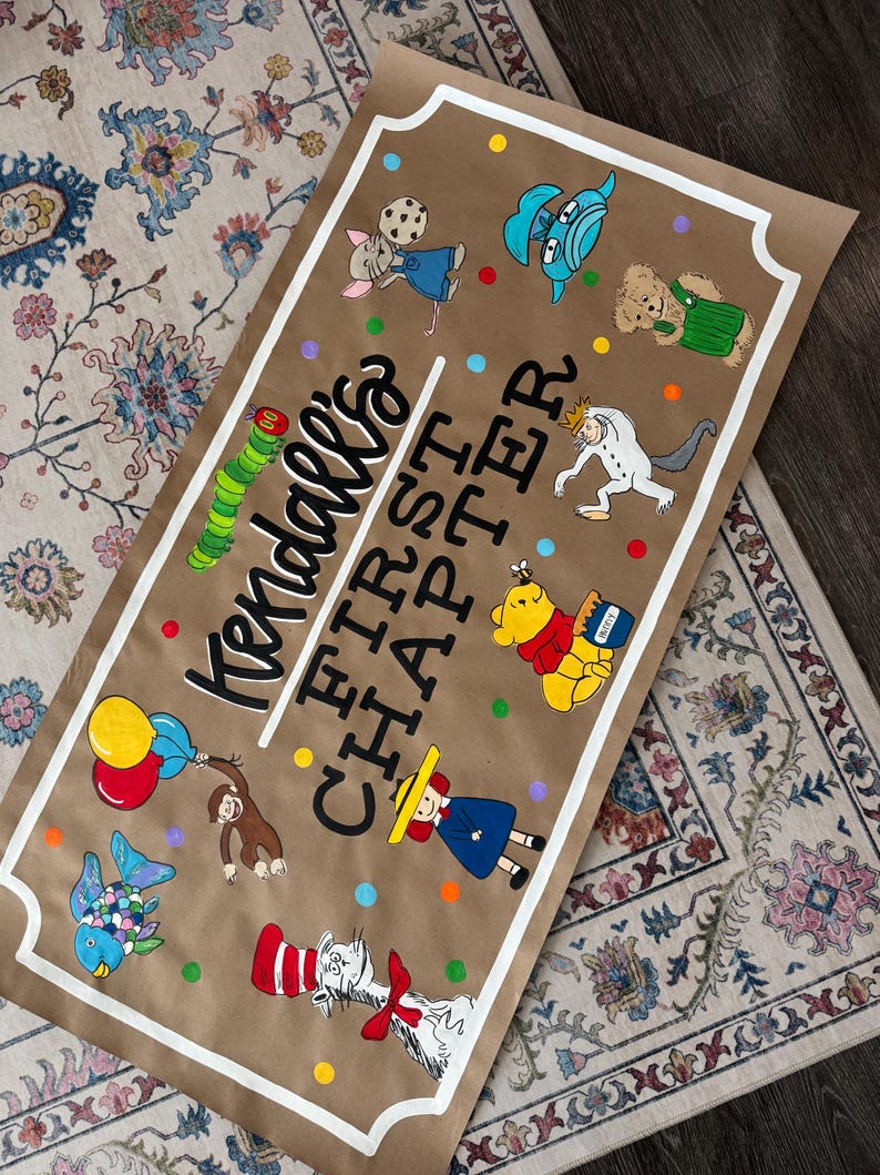 May include: A brown paper banner with the text "Kendall's FIRST CHAPTER" in black and white. The banner features colorful illustrations of storybook characters, including a mouse, a caterpillar, and Winnie the Pooh. The banner is trimmed with a white border.