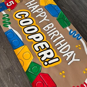 Pu&ograve; includere: Un banner di compleanno di carta marrone con le parole "HAPPY BIRTHDAY COOPER!" in bianco e giallo. Il banner &egrave; decorato con blocchi giocattolo colorati, una figura Lego e un palloncino verde.