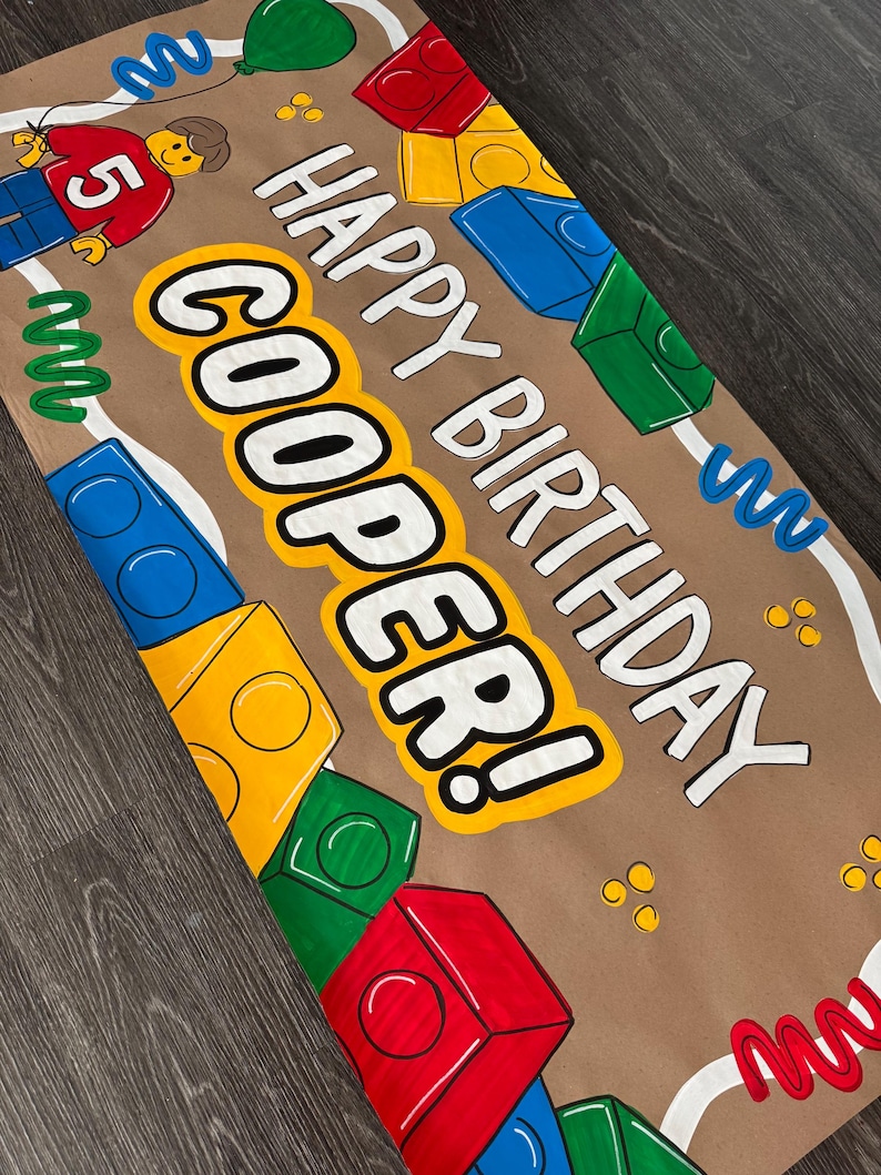 Pu&ograve; includere: Un banner di compleanno di carta marrone con le parole "HAPPY BIRTHDAY COOPER!" in bianco e giallo. Il banner &egrave; decorato con blocchi giocattolo colorati, una figura Lego e un palloncino verde.