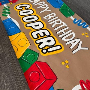 Pu&ograve; includere: Uno striscione di carta marrone con le parole "HAPPY BIRTHDAY COOPER!" in bianco e giallo, decorato con illustrazioni di blocchi colorati e una figura in stile Lego. Il numero "5" &egrave; visibile sulla maglietta della figura.