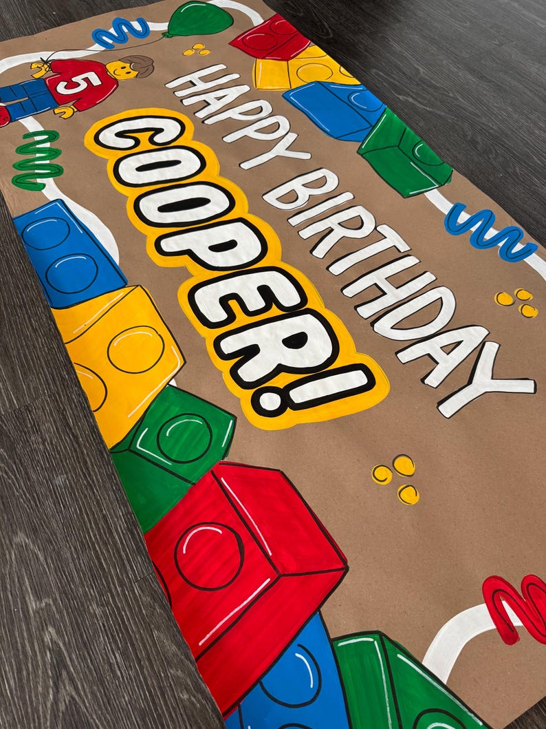 Pu&ograve; includere: Uno striscione di carta marrone con le parole "HAPPY BIRTHDAY COOPER!" in bianco e giallo, decorato con illustrazioni di blocchi colorati e una figura in stile Lego. Il numero "5" &egrave; visibile sulla maglietta della figura.