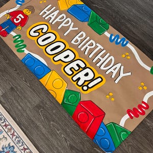 Pu&ograve; includere: Uno striscione di compleanno di carta marrone con le parole "HAPPY BIRTHDAY COOPER!" in lettere a blocchi bianche e gialle. Lo striscione presenta illustrazioni di blocchi colorati, una figura in stile Lego con il numero 5 e vortici decorativi.