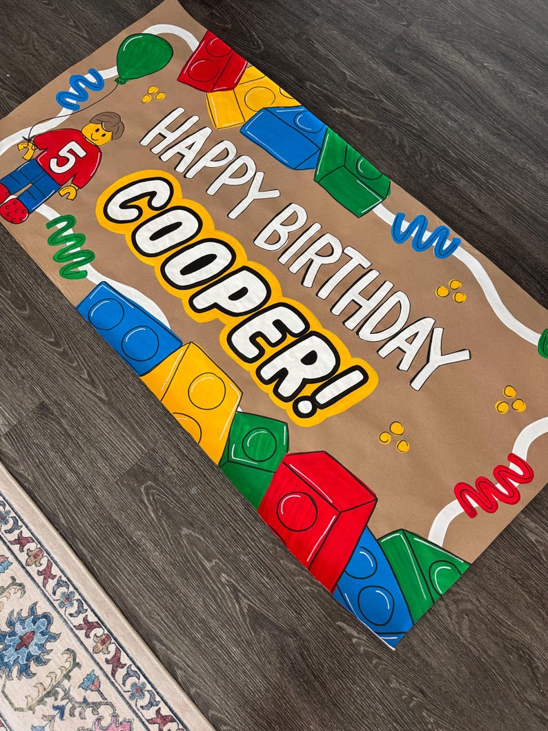 Pu&ograve; includere: Uno striscione di compleanno di carta marrone con le parole "HAPPY BIRTHDAY COOPER!" in lettere a blocchi bianche e gialle. Lo striscione presenta illustrazioni di blocchi colorati, una figura in stile Lego con il numero 5 e vortici decorativi.