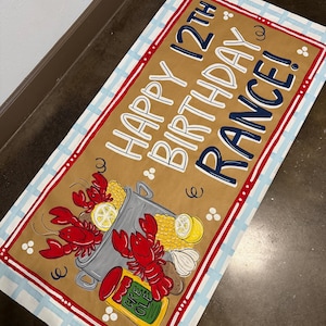 May include: A rectangular mat with a brown background and a blue and white striped border. The mat features a crawfish boil design with red crawfish, corn, lemons, and the text "HAPPY 12TH BIRTHDAY RANCE!"