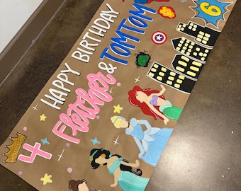 Può includere: Un banner di compleanno di carta marrone con le parole "Happy Birthday Fletcher & TomTom" in bianco, rosa e blu. Il banner presenta illustrazioni di supereroi, principesse e i numeri 4 e 6.