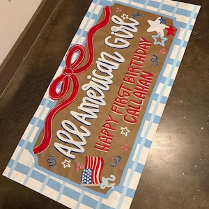 May include: A rectangular sign with a brown background and a blue and white checkered border. The sign reads "All American Girl" in white script with a red ribbon design. Additional text includes "Happy First Birthday Callahan" with star and flag graphics.