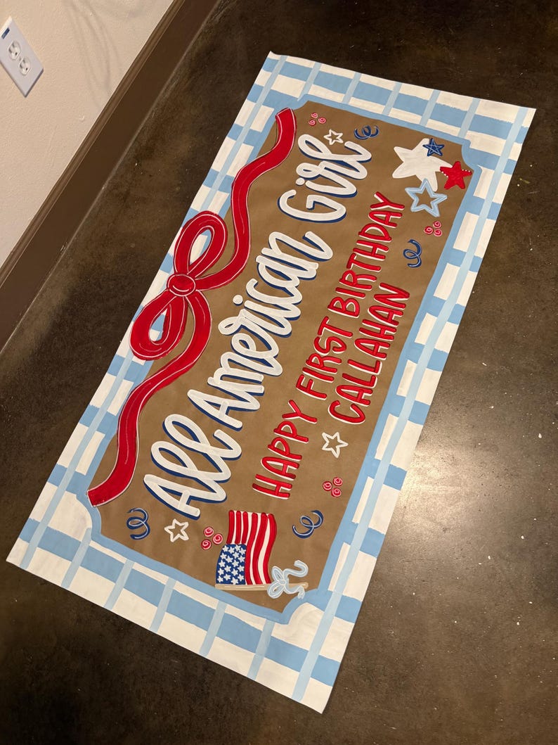 May include: A rectangular sign with a brown background and a blue and white checkered border. The sign reads "All American Girl" in white script with a red ribbon design. Additional text includes "Happy First Birthday Callahan" with star and flag graphics.