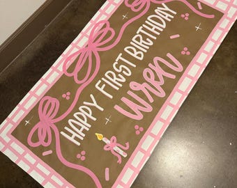 May include: A rectangular banner with the text "HAPPY FIRST BIRTHDAY Wren" in white and pink. The banner has a brown background with pink bows and a pink and white checkered border. A candle is also featured.