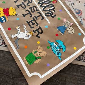 May include: Brown paper banner with the text "Madale's FIRST Easter" in black script. The banner features cartoon illustrations of Winnie the Pooh, a caterpillar, a mouse, and other characters. Colorful polka dots are scattered around the illustrations.