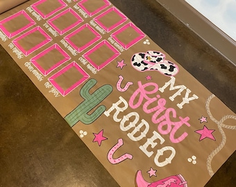 Może przedstawiać: Zwinięty brązowy papierowy plakat milowy z różowymi i białymi literami i ilustracjami. Tekst brzmi "My First Rodeo". Zawiera kaktus, kapelusz kowbojski, podkowę i but. Pola dla miesięcznych kamieni milowych są obrysowane na różowo.