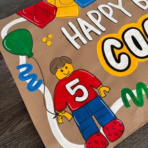 Pu&ograve; includere: Biglietto di compleanno dipinto a mano con un personaggio in stile Lego con una maglietta rossa con il numero 5, pantaloni blu e scarpe rosse. Il biglietto dice "HAPPY BIRTHDAY" e include un palloncino verde.