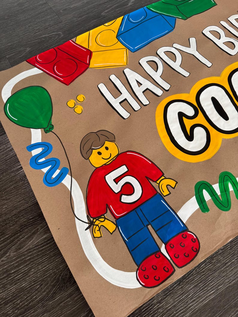Pu&ograve; includere: Biglietto di compleanno dipinto a mano con un personaggio in stile Lego con una maglietta rossa con il numero 5, pantaloni blu e scarpe rosse. Il biglietto dice "HAPPY BIRTHDAY" e include un palloncino verde.