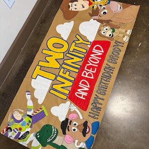May include: A brown banner featuring characters from the Toy Story movie. The banner includes Woody, Buzz Lightyear, Mr. Potato Head, Rex, and Slinky Dog. Text on the banner reads "Two Infinity and Beyond" and "Happy Birthday Brody!"