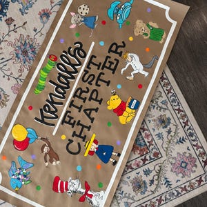 May include: A brown paper banner with the text "Kendall's FIRST CHAPTER" in black and white, surrounded by colorful cartoon characters. The banner is bordered with a white frame and features illustrations of a caterpillar, Winnie the Pooh, and other storybook characters.
