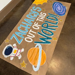 May include: A brown paper banner with the text "Zacharys Out of This World!" in blue and green lettering. The banner is decorated with a cartoon astronaut, a planet with rings, stars, and a swirling galaxy.