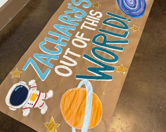 Può includere: Uno striscione di carta marrone con il testo "Zacharys Out of This World!" in lettere blu e verdi. Lo striscione è decorato con un astronauta dei cartoni animati, un pianeta con anelli, stelle e una galassia vorticosa.