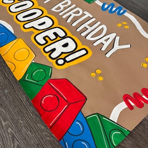 Pu&ograve; includere: Carta da regalo marrone con la scritta "HAPPY BIRTHDAY COOPER!" in bianco e giallo. Blocchi di costruzione in stile cartone animato colorati in rosso, blu, verde e giallo decorano la carta, insieme a zig-zag bianchi e blu.