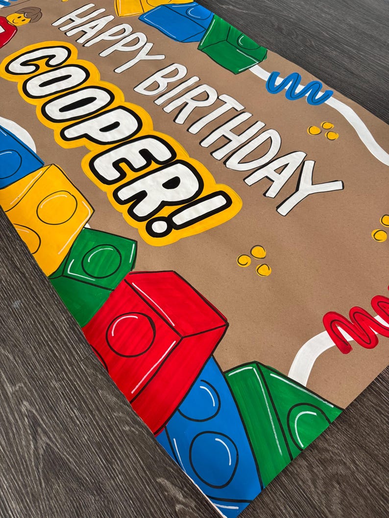 Pu&ograve; includere: Carta da regalo marrone con la scritta "HAPPY BIRTHDAY COOPER!" in bianco e giallo. Blocchi di costruzione in stile cartone animato colorati in rosso, blu, verde e giallo decorano la carta, insieme a zig-zag bianchi e blu.