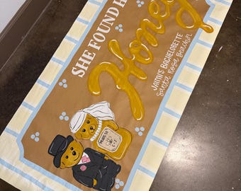 Può includere: Un tessuto rettangolare color crema con un pannello centrale marrone. Presenta le parole "SHE FOUND HER" e "Honey" in un carattere giocoso. Il design include orsi dei cartoni animati vestiti da sposa e sposo, con il testo "Jaimi's Bachelorette Santa Rosa Beach FL".