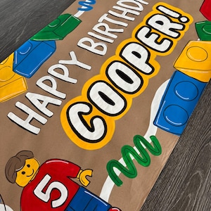 Pu&ograve; includere: Carta da regalo marrone con la scritta "HAPPY BIRTHDAY COOPER!" in bianco e giallo. Il design include blocchi colorati e una figura in stile Lego con una maglietta rossa con il numero "5".