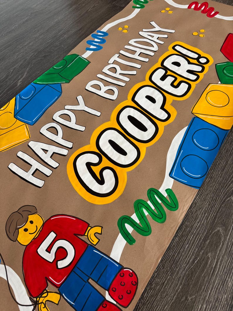 Pu&ograve; includere: Carta da regalo marrone con la scritta "HAPPY BIRTHDAY COOPER!" in bianco e giallo. Il design include blocchi colorati e una figura in stile Lego con una maglietta rossa con il numero "5".