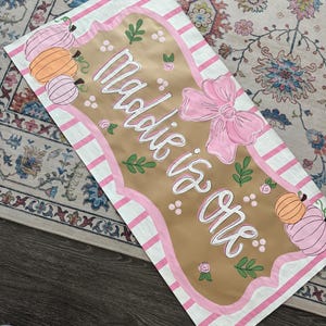 May include: A rectangular banner with the text "Maddie is One" in white script. The banner has a tan center with pink and white striped borders. Pink pumpkins and a pink bow decorate the banner, which is likely for a first birthday celebration.