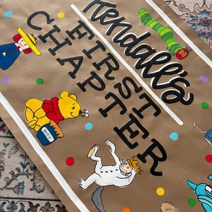 May include: A brown paper banner with the text "Kendall's FIRST CHAPTER" in black script and block letters. The banner features illustrations of cartoon characters, including Winnie the Pooh, Curious George, and the Cat in the Hat.