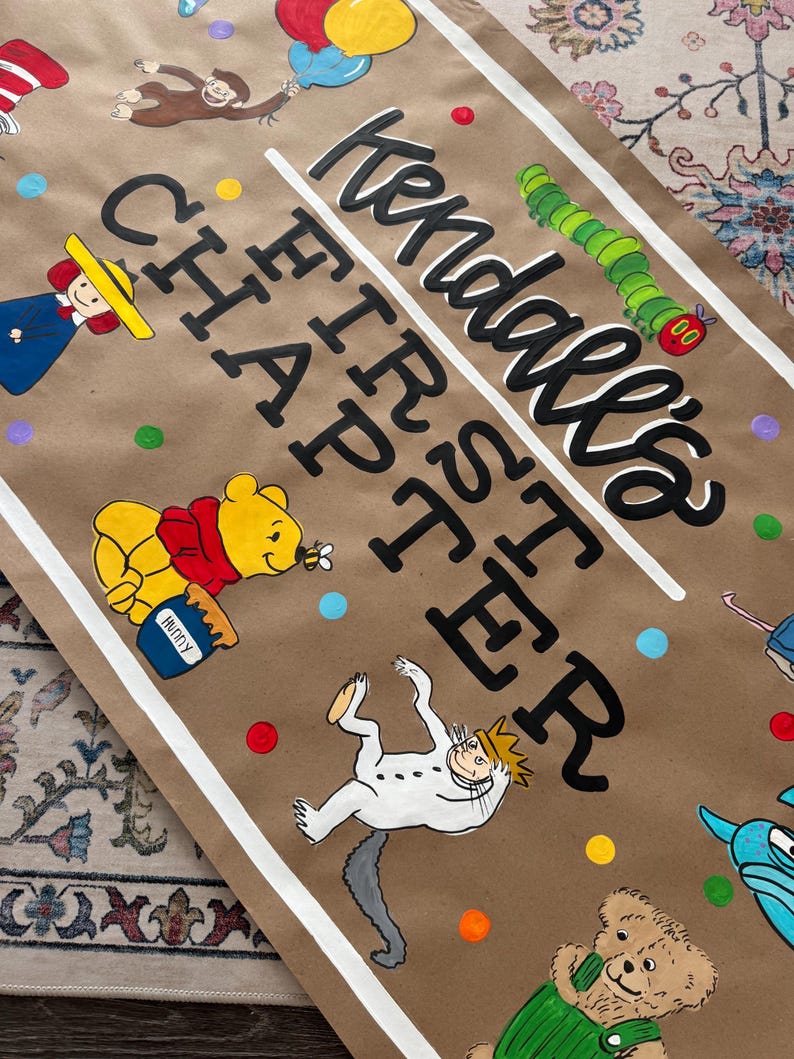 May include: A brown paper banner with the text "Kendall's FIRST CHAPTER" in black script and block letters. The banner features illustrations of cartoon characters, including Winnie the Pooh, Curious George, and the Cat in the Hat.