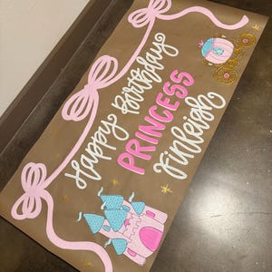 May include: A brown paper banner with the text "Happy Birthday PRINCESS Finleigh" in white and pink. The banner features pink bows, a pink and blue castle, a pink carriage, and gold star accents. Perfect for a princess-themed birthday party.
