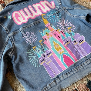 May include: A denim jacket with a hand-painted design of a pink and purple castle with fireworks. The name "Quinn" is painted in pink letters on the back of the jacket.