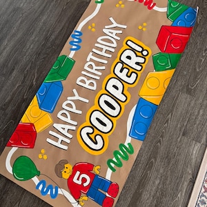 Pu&ograve; includere: Uno striscione di carta marrone con le parole "HAPPY BIRTHAY COOPER!" in bianco e giallo. Lo striscione presenta illustrazioni di blocchi colorati, un palloncino verde e una figura Lego con il numero 5. Sono presenti anche linee ondulate e punti decorativi.