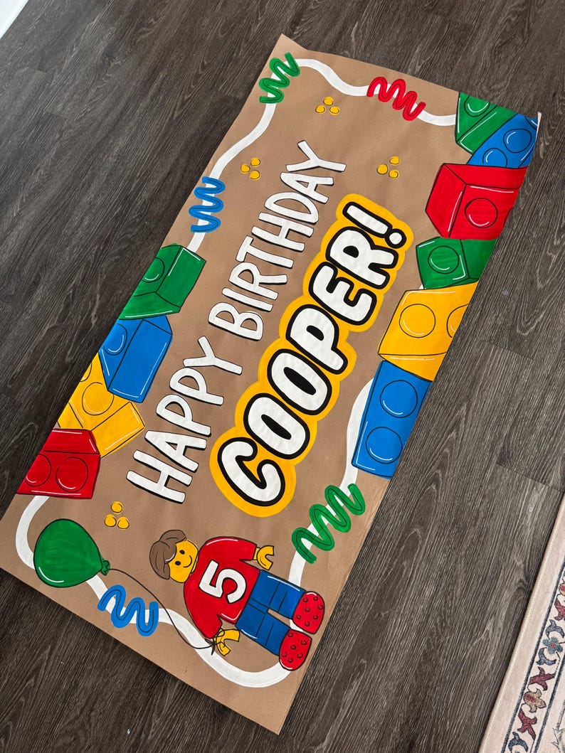 Pu&ograve; includere: Uno striscione di carta marrone con le parole "HAPPY BIRTHAY COOPER!" in bianco e giallo. Lo striscione presenta illustrazioni di blocchi colorati, un palloncino verde e una figura Lego con il numero 5. Sono presenti anche linee ondulate e punti decorativi.