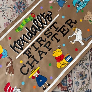 May include: A brown paper sign with the text "Kendall's FIRST CHAPTER" in black and white. The sign is decorated with colorful illustrations of children's book characters, including The Very Hungry Caterpillar and Winnie the Pooh.