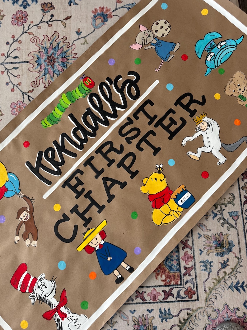 May include: A brown paper sign with the text "Kendall's FIRST CHAPTER" in black and white. The sign is decorated with colorful illustrations of children's book characters, including The Very Hungry Caterpillar and Winnie the Pooh.