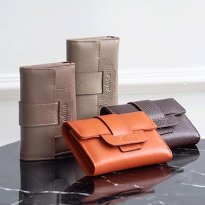 Può includere: Una collezione di portafogli in pelle in vari colori, tra cui marrone, beige e arancione. I portafogli hanno una finitura strutturata e presentano una chiusura a patta con fibbia. Il marchio "alona" è sottilmente impresso su ogni portafoglio.